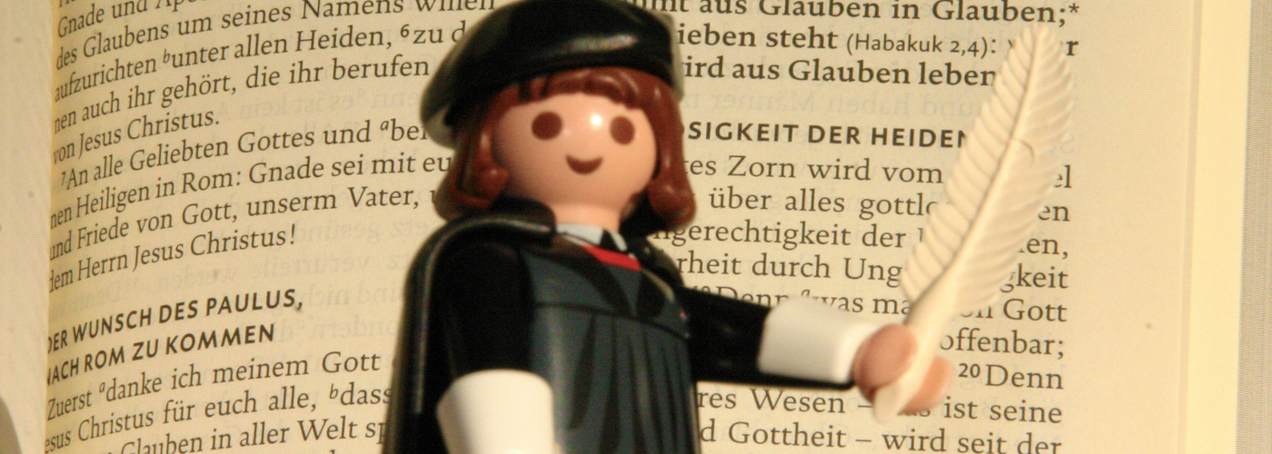 Das Bild zeigt Martin Luther als Playmobilfigur mit Feder und Bibel in der Hand, im Hintergrund der Römerbrief.